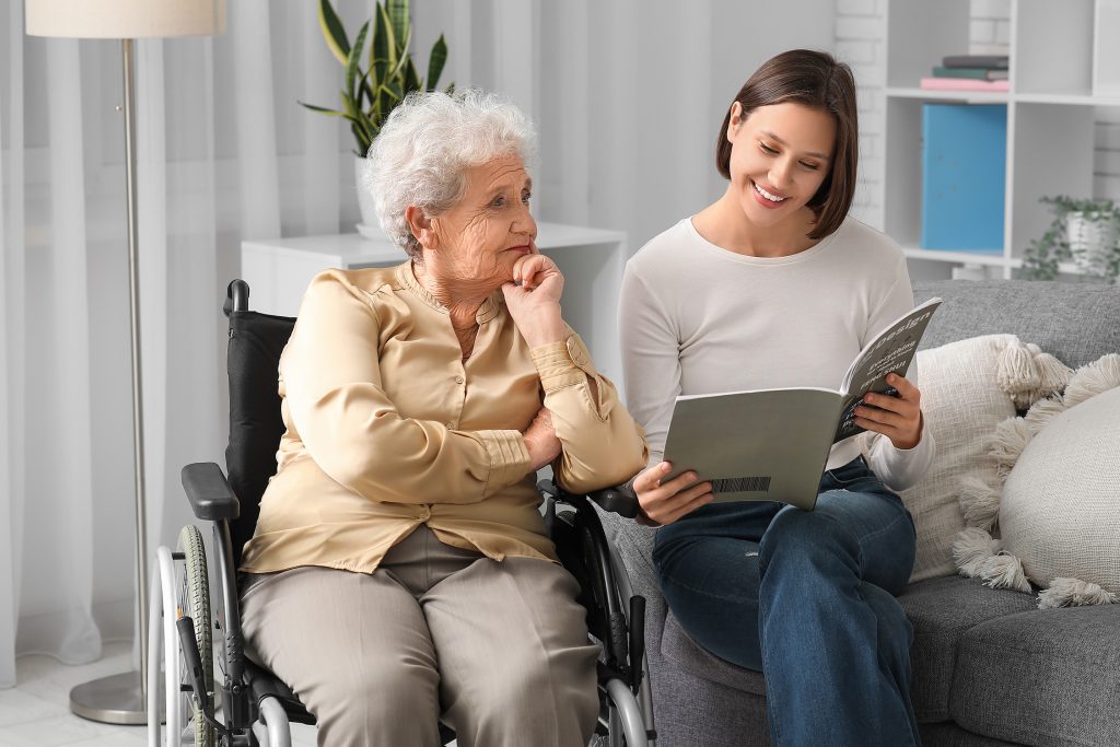 Betreuungskraft liest einer älteren Dame im Rollstuhl zu Hause aus einer Zeitschrift vor